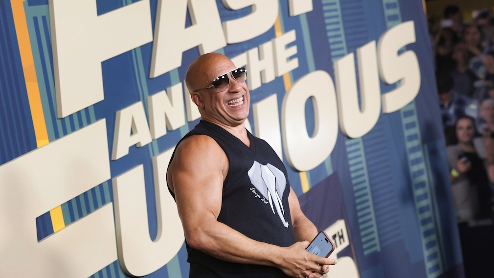 Vin Diesel steht vor einer großformatigen blauen Werbewand mit dem Schriftzug "FAST X" und weiteren großen Buchstaben. Die Person trägt eine dunkle ärmellose Oberbekleidung und eine Sonnenbrille und hält ein Smartphone in den Händen. Im Hintergrund sind unscharf weitere Menschen sowie Kameras zu sehen. Die Aufnahme zeigt einen Bereich einer Veranstaltung oder Premiere mit großflächiger Filmwerbung.