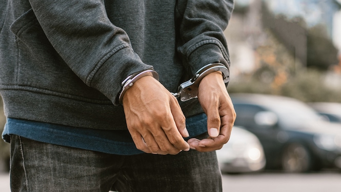 Nahaufnahme einer Person in dunklem Kapuzenpullover und Jeans, deren Hände vor dem Körper mit Metallhandschellen gefesselt sind. Die Arme hängen nach unten. Im unscharfen Hintergrund sind mehrere geparkte Fahrzeuge und eine städtische Umgebung zu erkennen.