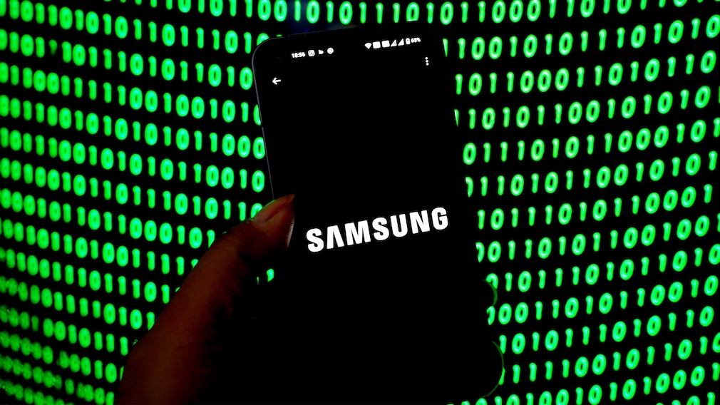 Eine Hand hält ein Smartphone im Hochformat vor einem Hintergrund aus leuchtend grünen Binärzahlen auf schwarzem Grund. Auf dem Display des Smartphones ist mittig das weiße Samsung-Logo auf schwarzem Hintergrund zu sehen. Oben auf dem Bildschirm sind verschiedene Statussymbole und ein Akkustand von 65 Prozent sichtbar. Die grünen Zahlen füllen den gesamten Hintergrund und sind unscharf dargestellt, während das Smartphone im Vordergrund scharf zu sehen ist.
