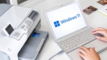Weißer Drucker und silberfarbenes Notebook auf einem Schreibtisch. Auf dem Bildschirm des Notebooks ist das Windows-11-Logo zu sehen. Eine Person bedient die Tastatur mit beiden Händen. Das Notebook ist per Kabel verbunden. Im Hintergrund liegen mehrere kleine Fotos auf der Tischoberfläche.