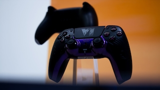 Schwarzer PlayStation-Controller mit violetten Akzenten auf einem transparenten Ständer. Das Touchpad zeigt ein eingeprägtes „VI“-Symbol. Die Tasten, Analogsticks und das Steuerkreuz sind gut sichtbar. Im Hintergrund befindet sich unscharf ein weiterer schwarzer Controller. Die Aufnahme ist in warmem orangefarbenem Licht gehalten.