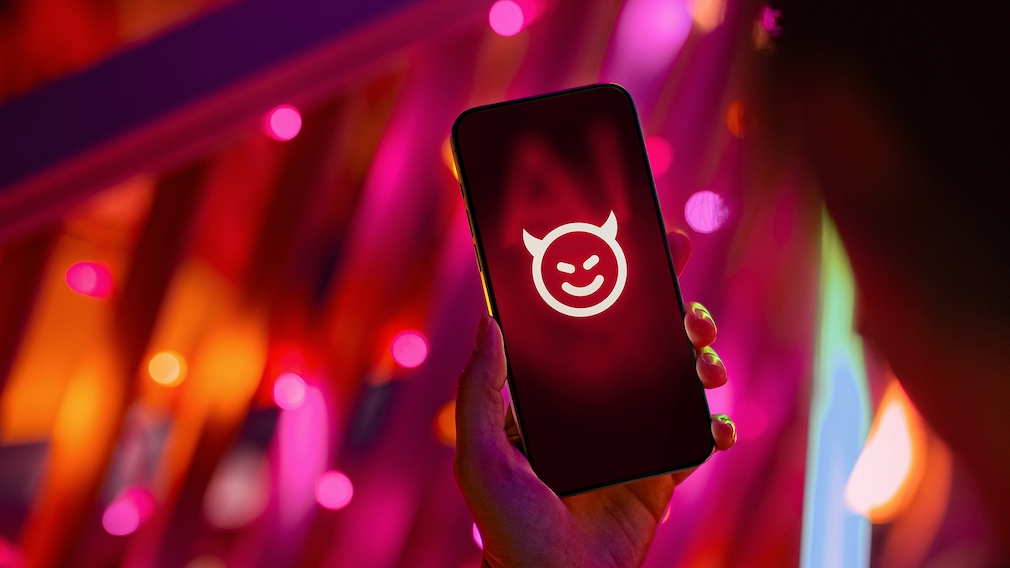 Eine Hand hält ein Smartphone vor einem unscharfen Hintergrund mit kräftigen roten, pinken und violetten Lichtquellen. Auf dem Display des Smartphones ist ein weißes Symbol zu sehen, das aus einem runden Gesicht mit Hörnern besteht. Das Telefon befindet sich zentral im Bild und wird von einer Hand am unteren Rand gehalten. Im Hintergrund sind farbige Lichtreflexe und verschwommene Formen erkennbar.