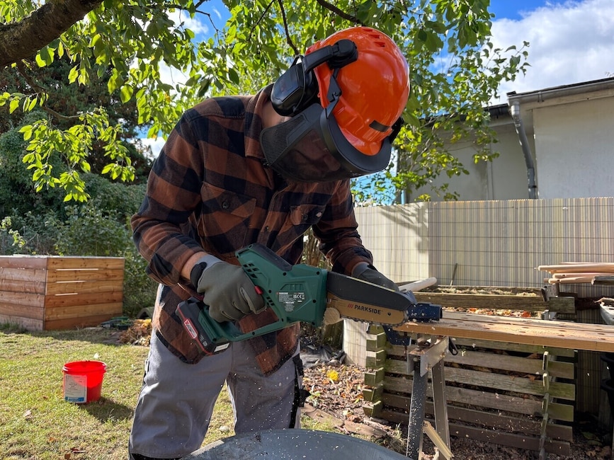 Ein Mann mit Schutzausrüstung sägt im Garten mit einer Mini-Kettensäge einen Besenstiel durch, der an einer Werkbank festgespannt ist. Im Hintergrund stehen Bäume und ein weißes Haus hinter einem Zaun.