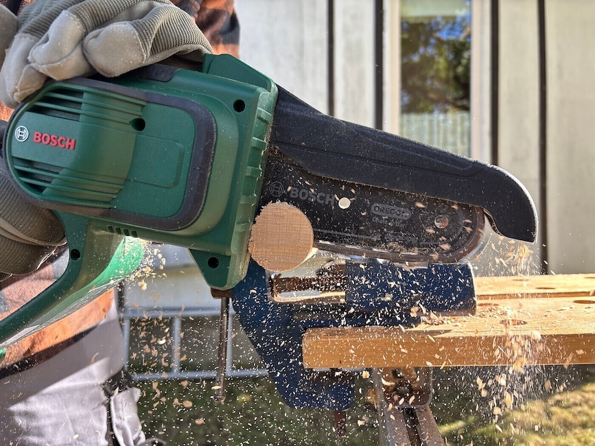 Eine Mini-Kettensäge von Bosch durchsägt ein Rundholz, das auf einer Werkbank festgespannt ist. Die Sägespäne fliegen.