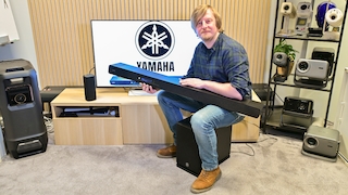Im Zentrum eines technisch ausgestatteten Wohnraums sitzt ein Mann auf dem schwarzen Subwoofer und hält die lange, flache Soundbar quer vor sich. Auf dem Fernseher im Hintergrund ist groß das Yamaha-Logo zu sehen; rundherum stehen weitere Lautsprecher, Projektoren und Unterhaltungselektronik. Helles Holz, schwarze Metallmöbel, ein heller Teppich und vertikale Holzlamellen verleihen der Szene einen modernen, wohnlichen Charakter.