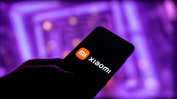 Eine Hand hält ein Smartphone mit Xiaomi-Logo und dem Schriftzug "Xiaomi" auf dem Display vor einem unscharfen, violett beleuchteten Hintergrund.