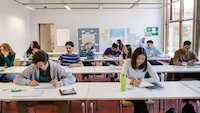Mehrere Schülerinnen und Schüler sitzen an Tischen in einem hellen Klassenzimmer. Einige schreiben in Hefte, andere unterhalten sich oder schauen auf ihre Smartphones.