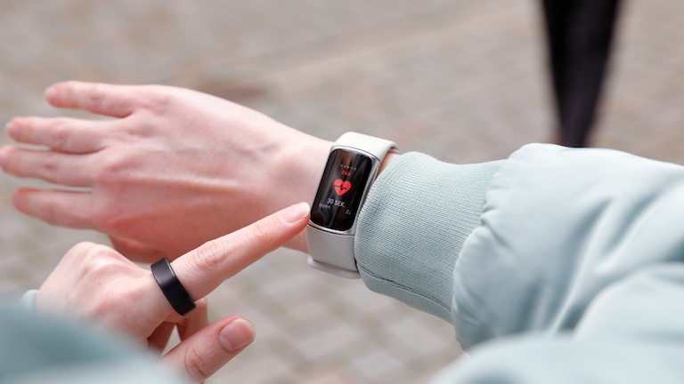 Nahaufnahme eines linken Arms mit einem Fitnessarmband mit weißem Armband und schmalem, hochkant ausgerichtetem Display. Die Anzeige zeigt eine EKG-Messung mit einem roten Herzsymbol und dem Text „EKG“ sowie „30 SEK“ und weiteren Angaben in kleiner Schrift. Eine rechte Hand mit schwarzem Ring am Zeigefinger berührt das Display. Die Person trägt eine hellgrüne Jacke. Im Hintergrund ist ein gepflasterter Boden sichtbar, leicht unscharf, sowie eine Person in dunkler Kleidung im oberen rechten Bildbereich.
